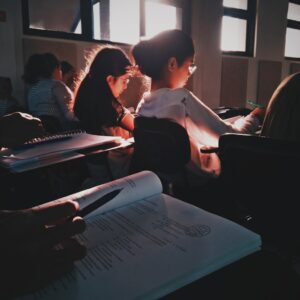 Gli studenti siedono ai banchi di un'aula, prendendo appunti e leggendo da libri aperti, con la luce del sole che filtra dalle finestre.