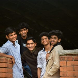 Cinque giovani uomini stanno vicini, sorridono e posano per una foto vicino a un muro di mattoni con una struttura scura sullo sfondo.