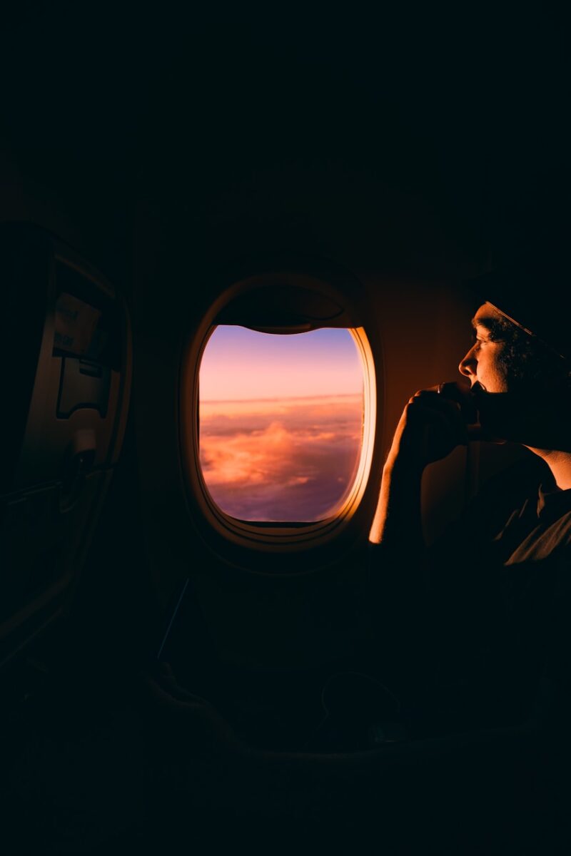 Una persona siede accanto al finestrino di un aereo al tramonto, guardando le nuvole rosa e arancioni illuminate dalla luce della sera, con un senso di nostalgia che cresce dentro di lei - giunta da lontano dopo un lungo viaggio.