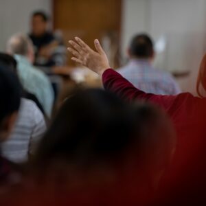 Persona che alza la mano in un contesto di gruppo, di fronte a un oratore nella parte anteriore della stanza, con diversi altri partecipanti seduti nelle vicinanze.